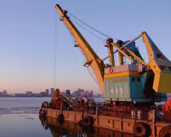 Grúa portuaria sobre una barcaza al atardecer, con la ciudad al fondo